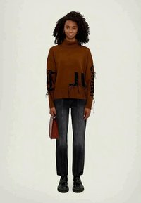 Rudas per didelės dydžio megztinis su juodais grafikų dizainais ir suplėšytais kraštais, derinamas su tamsiomis džinsomis ir juodais masyviais batais. Laiko rudą rankinę.