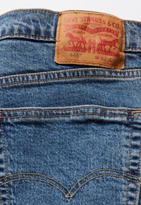 Džíny Levi's 445 s hnědou koženou etikou, na které je uvedeno "Levi Strauss & Co." v červené barvě, vyrobené z modré džínoviny, s oranžovým prošíváním a klasickými zadními kapsami.