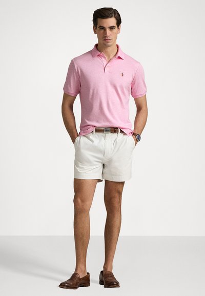 Polo de algodón en color rosa claro con cuello y pequeño logo, combinado con pantalones cortos blancos y mocasines de cuero marrón, llevando un cinturón marrón.