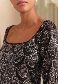 Čierny top zdobený striebornými flitrami v oblúkovom vzore, s širokým výstrihom a textúrovaným povrchom.