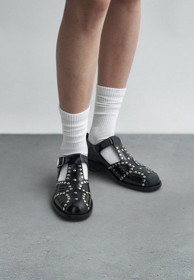 Sapatos de verniz preto com tachas prateadas, design em T e um pequeno salto bloco. Usados com meias brancas caneladas.