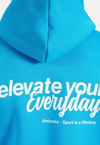 Un hoodie bleu clair avec un grand design en texte blanc au dos qui indique "élever votre quotidien" et "Smilodox - Le sport est un style de vie."