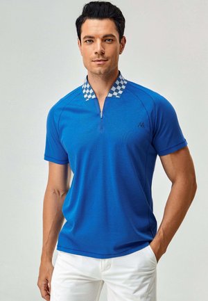 Uomo che indossa una polo blu a maniche corte con colletto a quadri e pantaloni bianchi, in piedi con una mano in tasca.