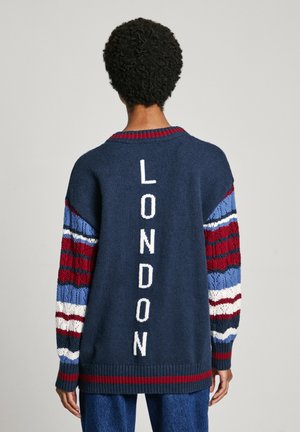Person med kort krøllet hår ifører sig en marineblå sweater med røde, hvide og blå stribede ærmer og teksten "LONDON" skrevet vertikalt på ryggen.