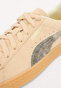 Beige velúr sportcipő fűzőkkel, amely texturált fekete csíkkal és gumitalppal rendelkezik. Az oldalán arany színben felületlenített logó található.