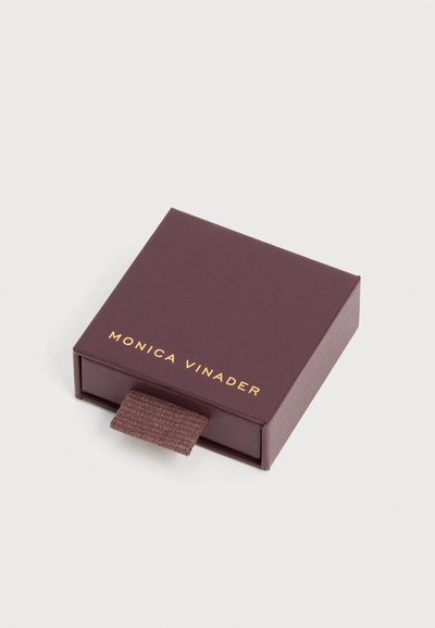 Τετράγωνο σκούρο μοβ κουτί κοσμημάτων με καφέ υφασμάτινη ταινία έλξης, με την επιγραφή "Monica Vinader" σε χρυσό στο καπάκι.