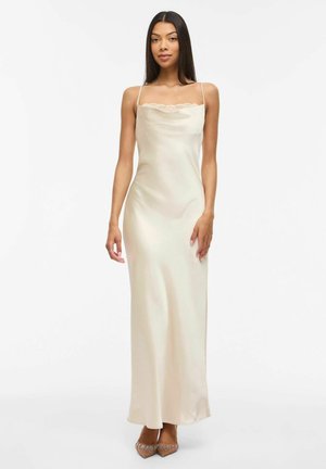 Femme aux longs cheveux noirs portant une robe nuisette en satin crème à fines bretelles et bordure en dentelle, associée à des talons pointus ornés.