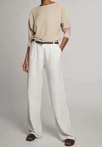 Top a maniche lunghe beige a costine, pantaloni bianchi a gamba larga e cintura marrone. L'insieme presenta un tessuto testurizzato e una vestibilità ampia, abbinato a sandali.