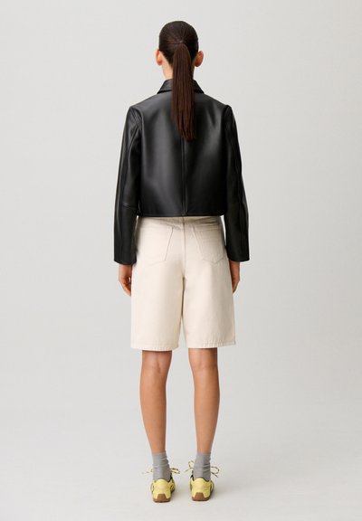 Mujer con coleta oscura que lleva una chaqueta de cuero negra, pantalones cortos beige hasta la rodilla, calcetines grises y zapatillas amarillas, de pie de espaldas.