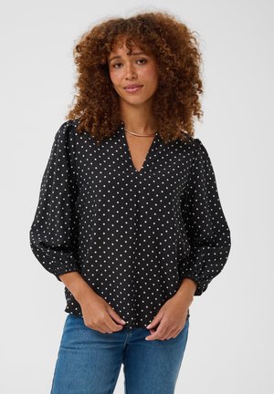 Femme aux cheveux bouclés portant un chemisier noir à pois blancs et un jean bleu, debout devant un fond clair uni.