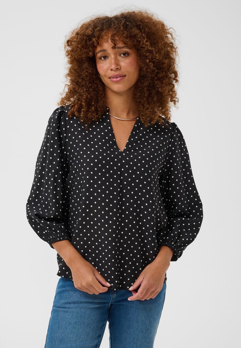 Femme aux cheveux bouclés portant un chemisier noir à pois blancs et un jean bleu, debout devant un fond clair uni.