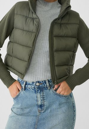Personne portant une veste matelassée sans manches vert olive par-dessus un pull gris côtelé et une jupe en denim bleu clair, mains dans les poches de la veste.