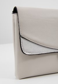 Petit sac pochette beige en cuir avec un détail de rabat triangulaire argenté métallique et une bordure noire, vu sous un angle.