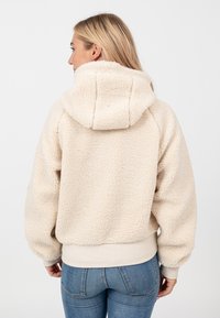 Kremowa polarowa bluza z kapturem o teksturowanym, pluszowym wykończeniu. Posiada duży kaptur i ribbowany dół. Widok z tyłu, luźny krój i szerokie rękawy.