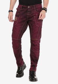 Bordeaux kleurige broeken van duurzaam materiaal, met decoratieve stiksels, meerdere zakken en een slim fit ontwerp. Draagt bij zwarte laarzen.