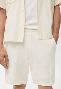 Shorts blancs en tricot avec une finition texturée, dotés de poches latérales et d'une taille élastique, portés avec une chemise à manches courtes de couleur claire.