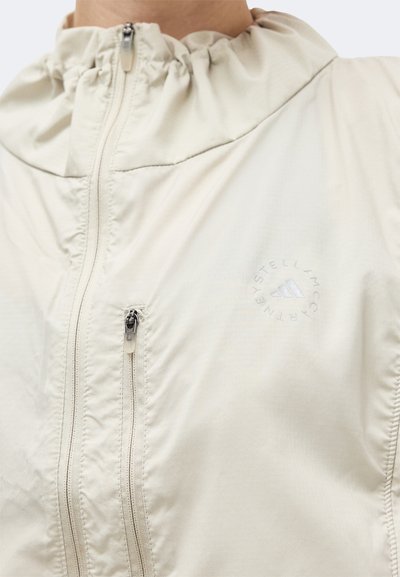 Μπεζ μπουφάν με φερμουάρ και ψηλό γιακά, με μικρή τσέπη με φερμουάρ στο στήθος και διακριτικά λογότυπα Stella McCartney και Adidas.