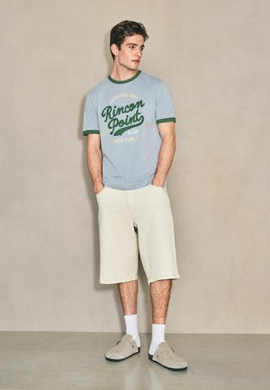 Junger Mann steht mit den Händen in den Taschen, trägt ein hellblaues "Rincon Point" T-Shirt, beige kniehohe Shorts, weiße Socken und beige Slipper.