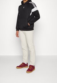 Black hooded sweatshirt with white side accents, kangaroo pocket, paired with light beige pants and maroon sneakers featuring yellow laces.