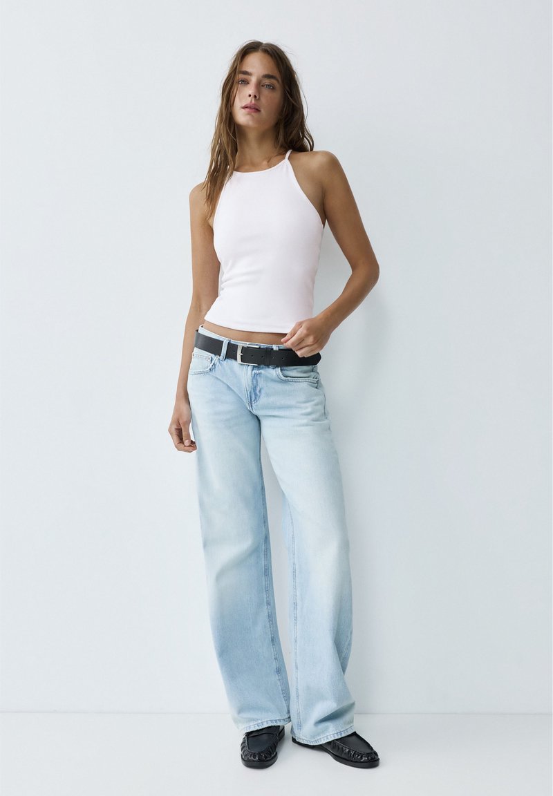 Jeune femme appuyée contre un mur blanc, portant un haut blanc sans manches, un jean bleu clair à jambes larges, ceinture noire et chaussures noires.