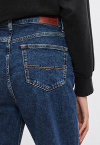 Tmavomodré džínsové nohavice majú hnedý kožený štítok na páse, zadné vrecká s oranžovým prešívaním a rovný strih.