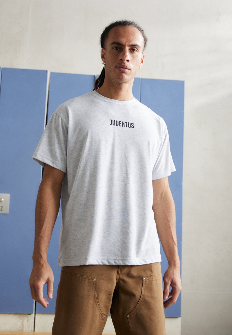 T-shirt de algodão cinza com mangas curtas, apresentando "JUVENTUS" impresso em preto no peito. O modelo também usa calças castanhas. Fundo texturizado.