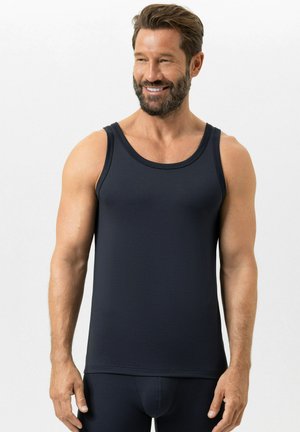 Uomo muscoloso con barba che indossa una canottiera nera aderente senza maniche e pantaloni abbinati, sorridente, in piedi davanti a uno sfondo bianco semplice.