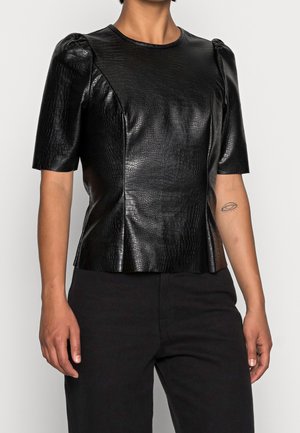 Top corto in finta pelle nera a maniche corte con un sottile motivo coccodrillo, spalle gonfie, indossato con pantaloni neri, tatuaggio sul braccio visibile.