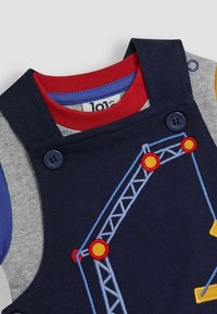 Salopette en coton bleu marine avec des accents gris, rouge et bleu. Présente un motif de broderie de grue en jaune, rouge et bleu. Fermeture par boutons.