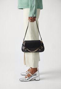 Černá kožená kabelka se zlatým řetízkovým detailem, s pevnou konstrukcí a plochým horním okrajem. Kombinováno se stříbrnými botami na blokovém podpatku.