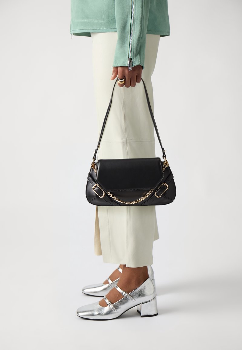 Černá kožená kabelka se zlatým řetízkovým detailem, s pevnou konstrukcí a plochým horním okrajem. Kombinováno se stříbrnými botami na blokovém podpatku.