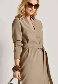 Beige lange mouw jurk met een V-hals en een riem rond de taille. Beschikt over een zachte stof, een ontspannen pasvorm en subtiele plooien. Accessoires omvatten gestapelde armbanden.