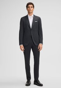 Completo grigio scuro con due bottoni, revers a coltello e tasche laterali. Un paio di scarpe in pelle nera, camicia bianca sotto la giacca.