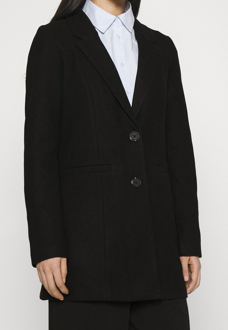 Persona che indossa un blazer nero a due bottoni sopra una camicia colletto blu chiaro, mostrata dalla gola ai fianchi, in piedi contro uno sfondo neutro.