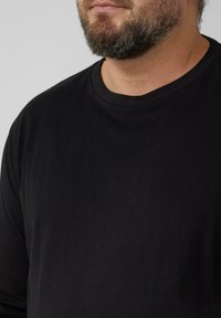 Bärtiger Mann mit einem schlichten schwarzen Langarm-Shirt mit Rundhalsausschnitt, dargestellt von Hals bis zur Brustmitte vor einfachem Hintergrund.