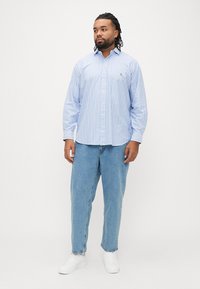 Camisa de manga comprida listrada azul-claro com gola com botões, usada com jeans de ganga soltos e ténis brancos. Estilo simples e casual.
