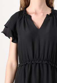 Black dress featuring short ruffled sleeves, a V-neckline, and an elasticized waist with ruffle detailing. Smooth, lightweight fabric.