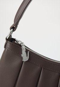 Sac à main en cuir marron avec une fermeture éclair argentée et un tirette en forme de crocodile sur une sangle rembourrée, sur fond blanc.