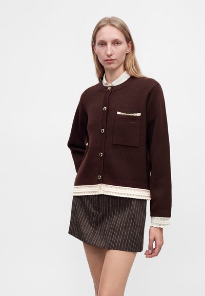 Femme portant un cardigan boutonné marron foncé avec une bordure en dentelle crème, par-dessus une chemise blanche, et une jupe sombre à fines rayures, sur un fond uni.
