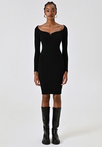Abito nero a coste con maniche lunghe e scollatura a cuore, caratterizzato da un dettaglio dorato al centro e una silhouette aderente.
