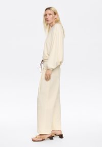 Maglione oversized beige chiaro con texture a coste, coulisse regolabile in vita, abbinato a pantaloni larghi beige chiaro e sandali con cinturini.