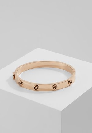 Armbånd i roseguld metal med jævnt placerede runde logonitter, hvilende på en hvid kube mod en grå baggrund.