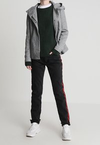 Grijze capuchonjas met knoopsluiting, groene geribbelde trui eronder, zwarte broek met rode zijstrepen en witte sneakers.