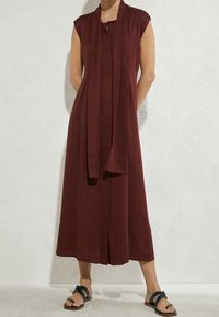 Ärmelloses, bordeauxfarbenes Midikleid mit Frontschlitz, kombiniert mit passendem Schal und schwarzen flachen Sandalen, getragen von einer Person vor einer weißen Wand.