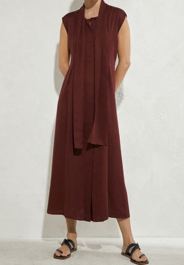 Ärmelloses, bordeauxfarbenes Midikleid mit Frontschlitz, kombiniert mit passendem Schal und schwarzen flachen Sandalen, getragen von einer Person vor einer weißen Wand.