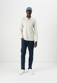 Camisa polo de manga larga en color beige claro con cuello, combinada con unos vaqueros azul oscuro y zapatillas blancas, complementada con una gorra azul claro.