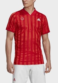 Camiseta deportiva roja con rayas verticales, de manga corta y cuello en V. Presenta el logo de Adidas en el pecho y una textura en la tela.