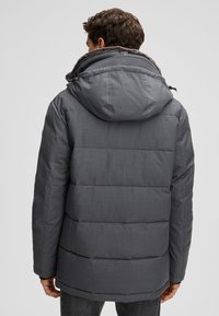 Mann trägt eine dunkelgraue gesteppte Winterjacke mit Kapuze, die mit braunem Kunstpelz gefüttert ist, von hinten gesehen.