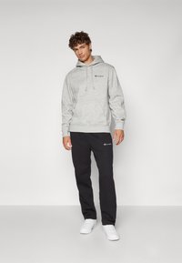 Grauer Baumwollhoodie mit Kordelzug und gesticktem Logo, kombiniert mit schwarzen Jogginghosen und weißen Turnschuhen, vor einem schlichten Hintergrund.