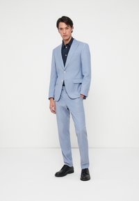 Paprastas mėlynas kostiumas su siaura siluetu, tekstūruota medžiaga, vienu mygtuku užsegamas švarkas ir atitinkančios kelnes. Derinamas su tamsia marškinėliais ir juodais batais.